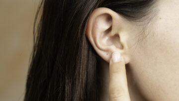 Ohren reinigen: Ein Zeigefinger zeigt auf das Ohr einer Frau mit braunen Haaren.